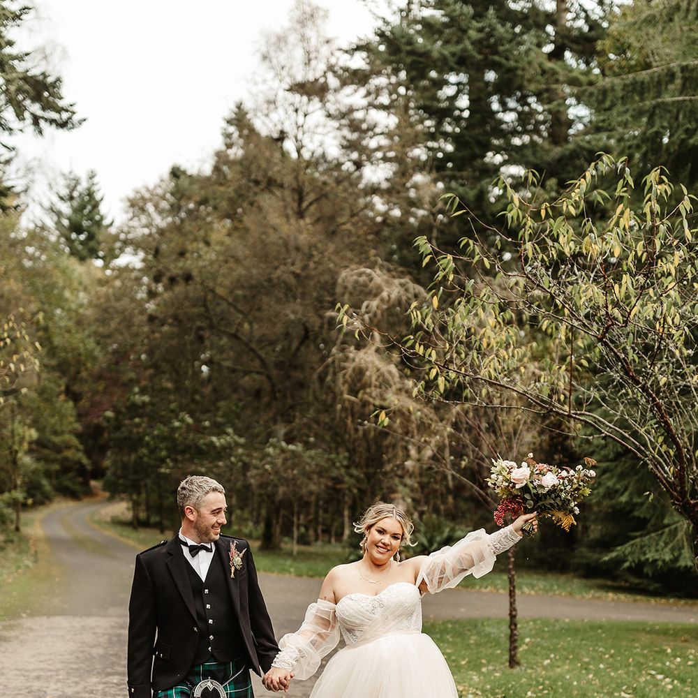 Groom in black tie and green kilt smiles at bride in custom gown lifting her bouquet into the air