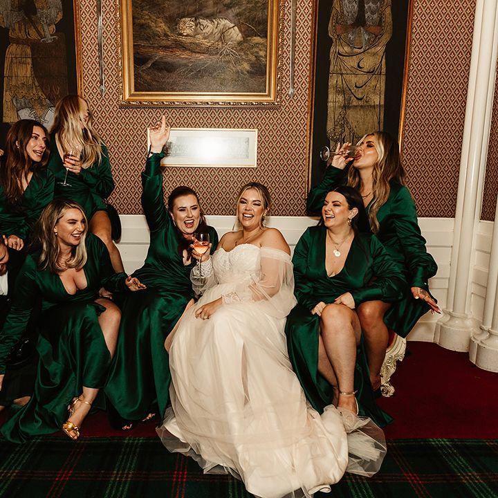 Bride sits holding glass of wine with bridesmaids in emerald green long sleeve dress with front slit also drinking