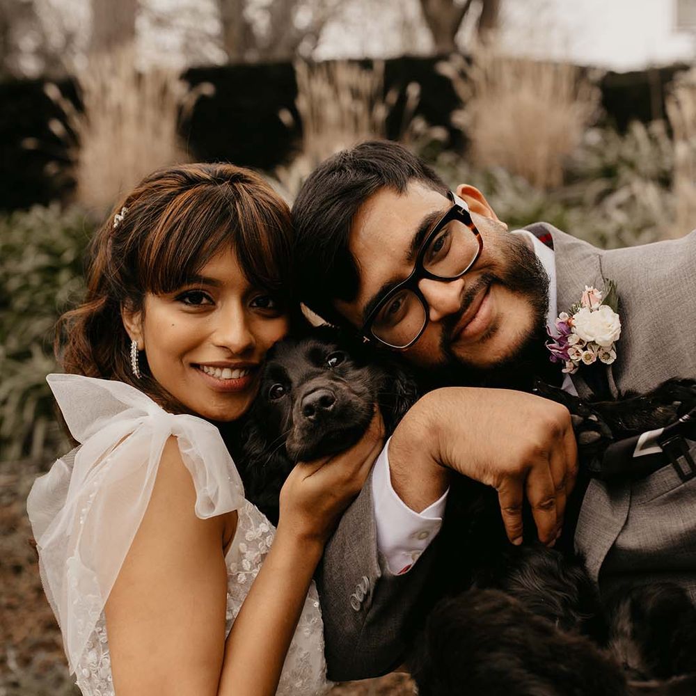 Bride smiling in white traditional wedding dress holding black puppy with groom, also smiling, in grey suit and bow tie