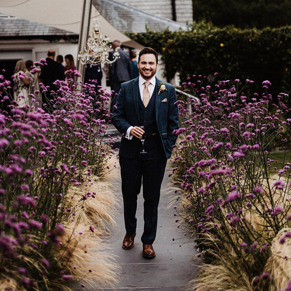 Groom stands next to purple wildflowers wearing blue suit and gold patterned tie and handkerchief