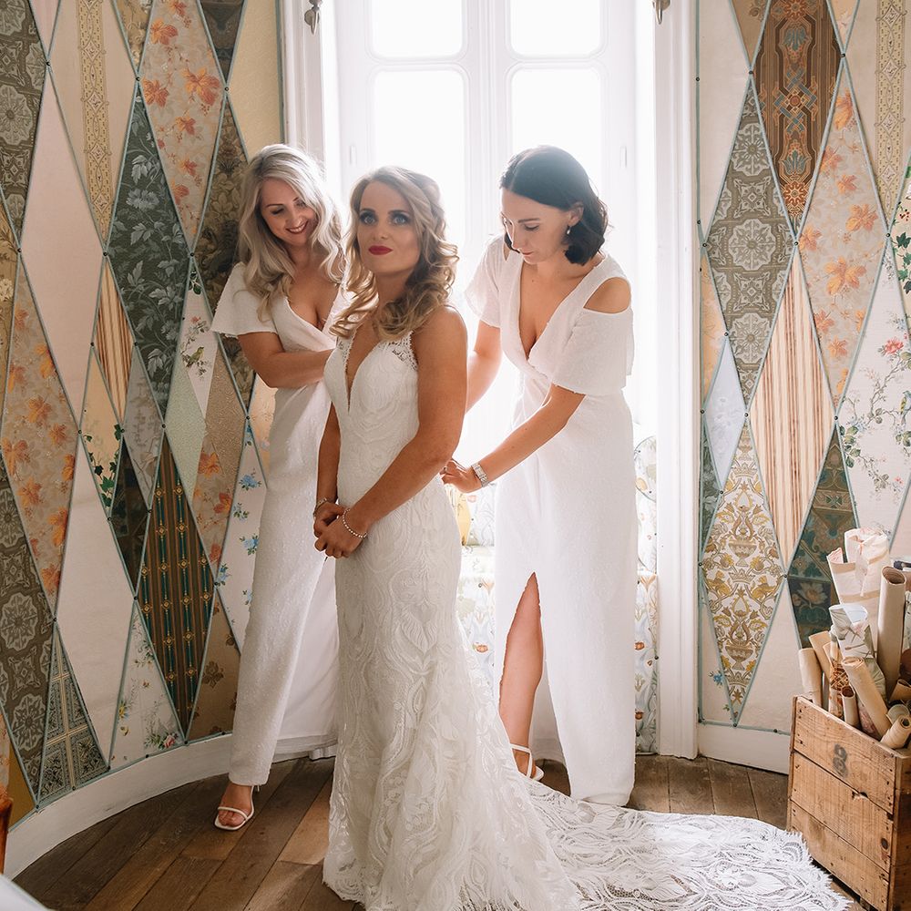 Bridesmaids in sparkly white dresses help the bride into her lace wedding dress with a long train with red lipstick and silver jewellery 