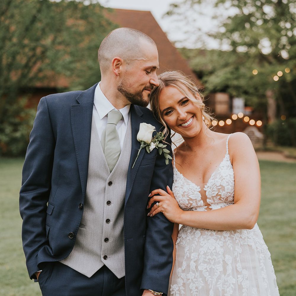 Groom in a dark blue suit with a grey waistcoat and green tie with a white rose buttonhole and the bride in a lace wedding dress take a cute couple portrait