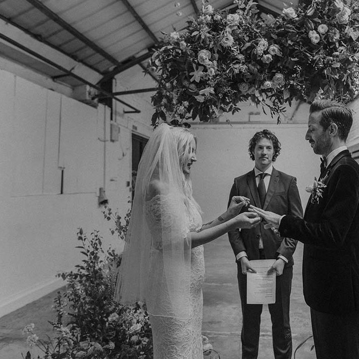 Bride in lace boho two piece with tattoos and groom in black tux with bow tie and boutonniere giving wedding rings at Hackney Studios