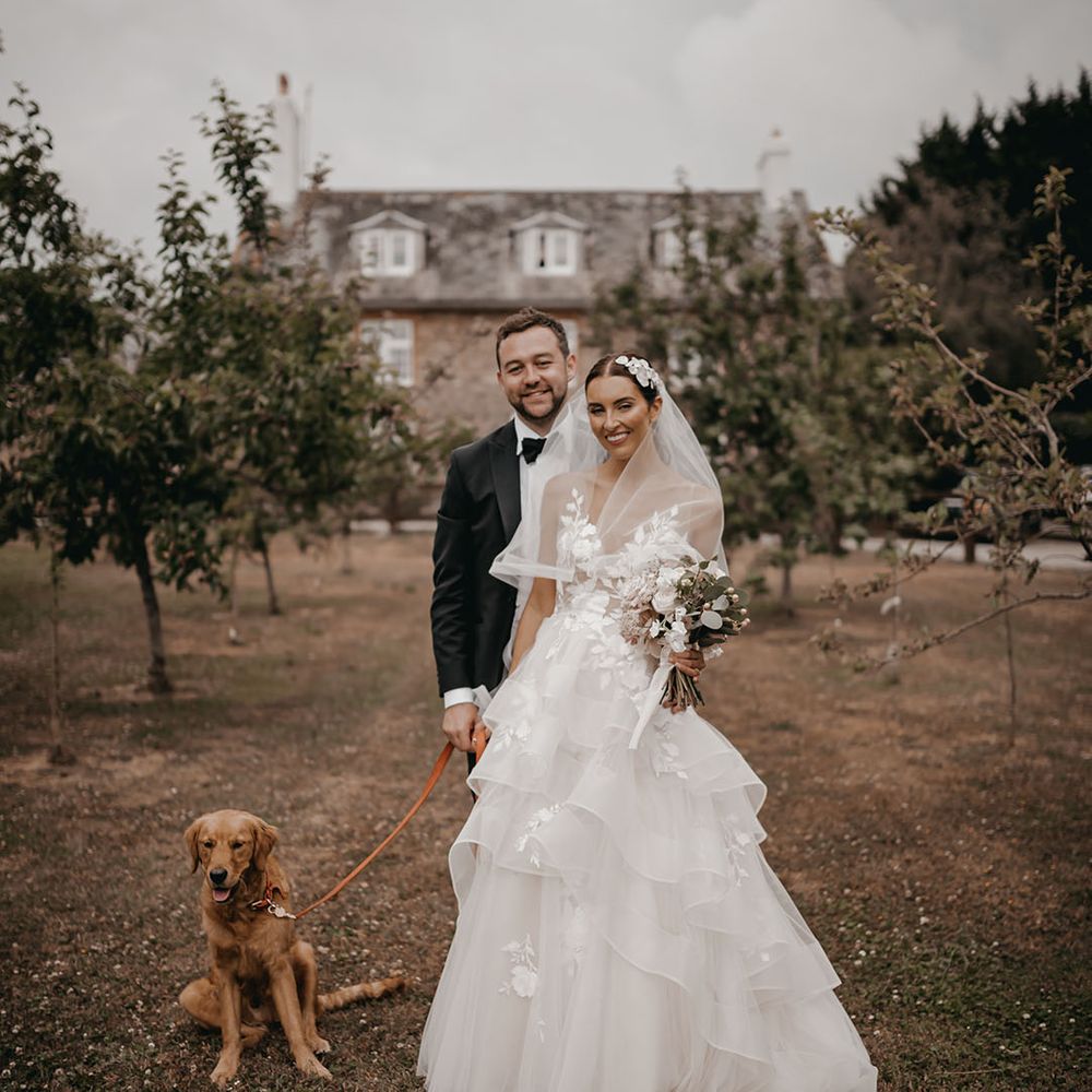 The bride and groom smile as they pose with their pet dog on a lead for their wedding at Anran