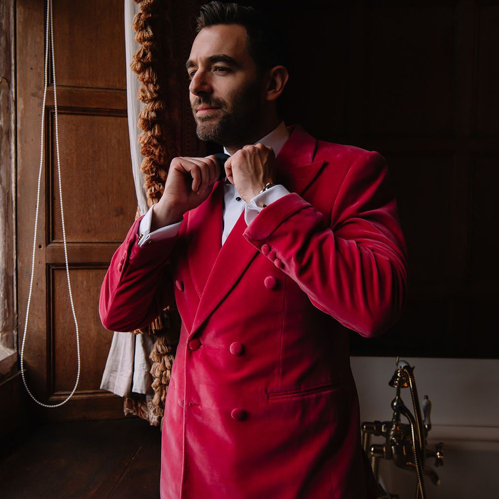 Groom puts on his black bow tie wearing a double breasted velvet suit jacket in a bright pink colour 