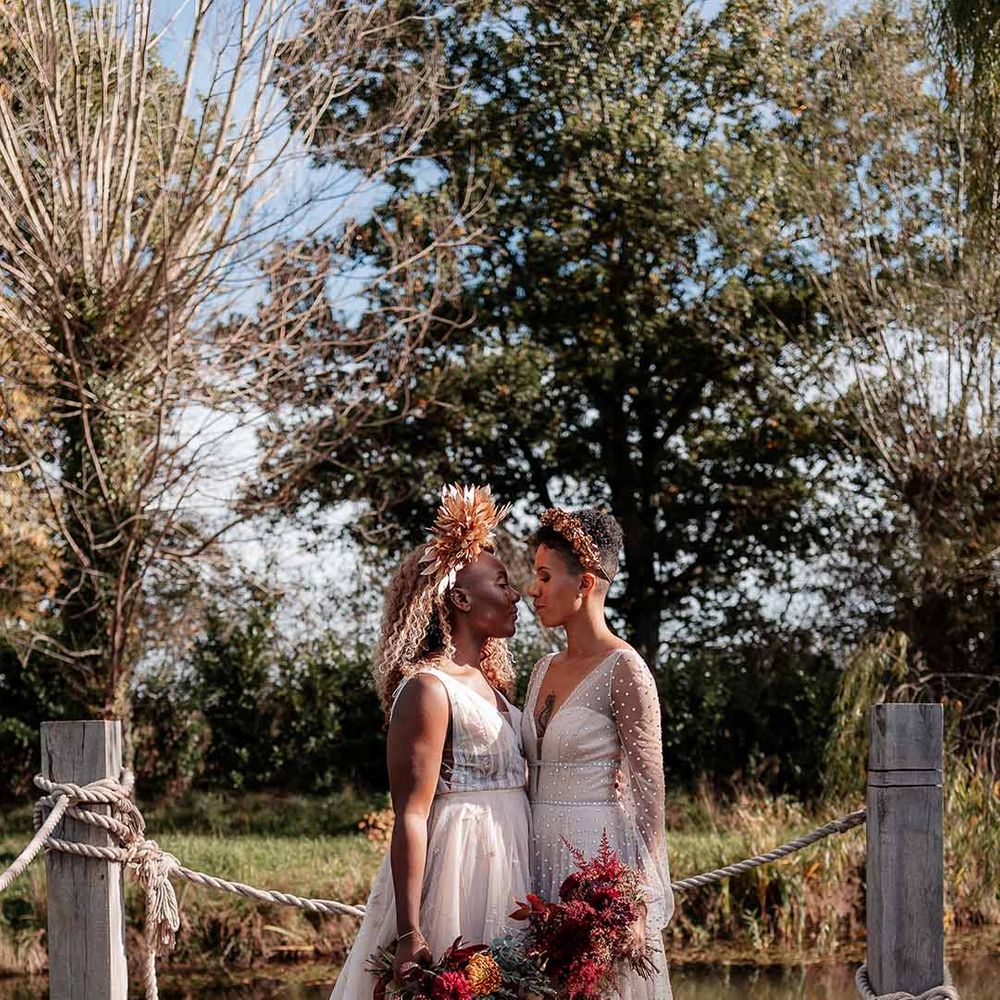 Bride in crystal embellished gown and gold headdress embracing bride in sleeveless floaty peach gown holding dried autumnal floral bouquets on a dock by the river at Silchester Farm