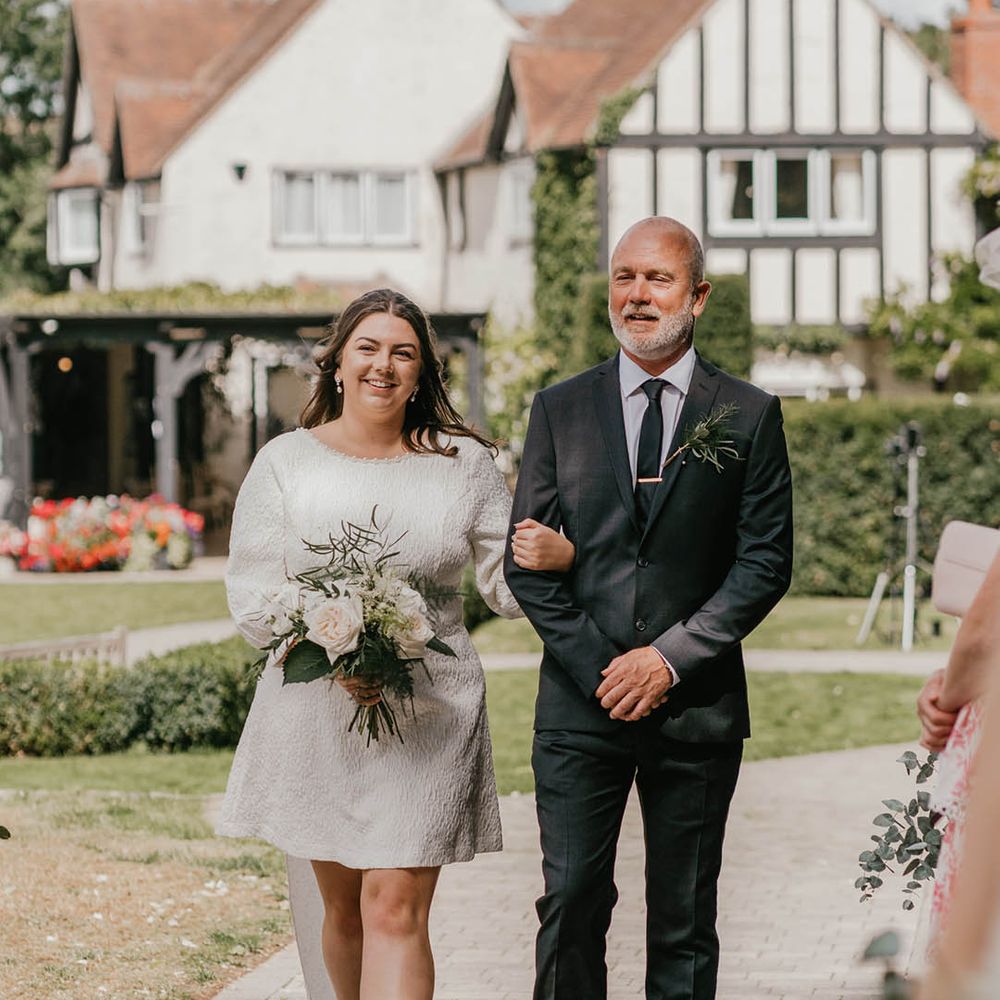 Father of the bride in a black suit and bride in a short wedding dress with bow detail holding all white wedding bouquet 