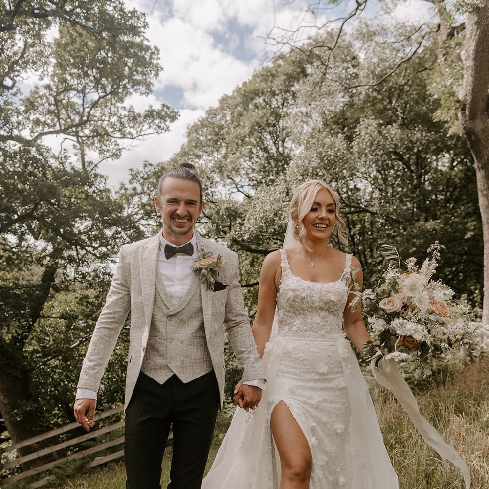 The bride in an Eleganza Sposa Iconica floral wedding dress with split leg walking along with the groom in a three piece light grey suit 