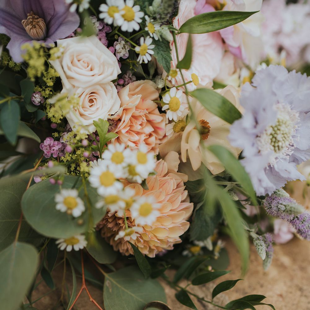 The bride's thin gold wedding band and groom's thicker gold wedding band rest together next to the bridal bouquet with pink, purple, and orange flowers