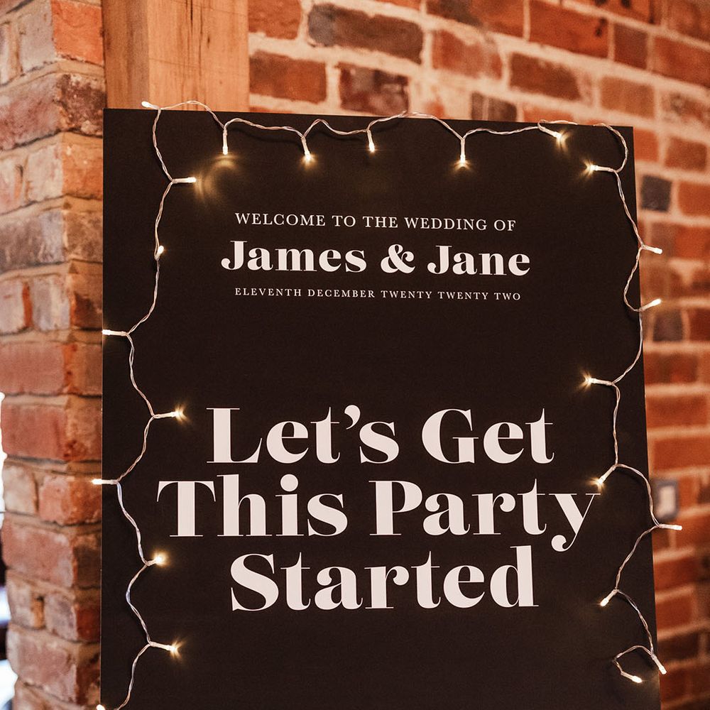 Black and white modern wedding welcome sign with fairy lights decorating the signage on a wooden easel 