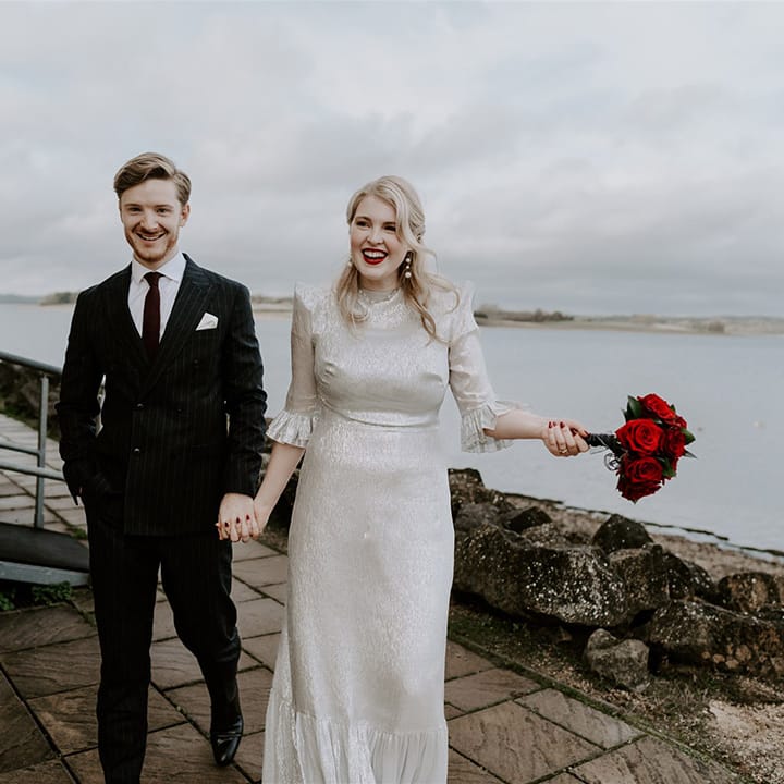 Groom in dark suit with red tie walking with bride in sparkly silver wedding dress holding red rose bouquet 