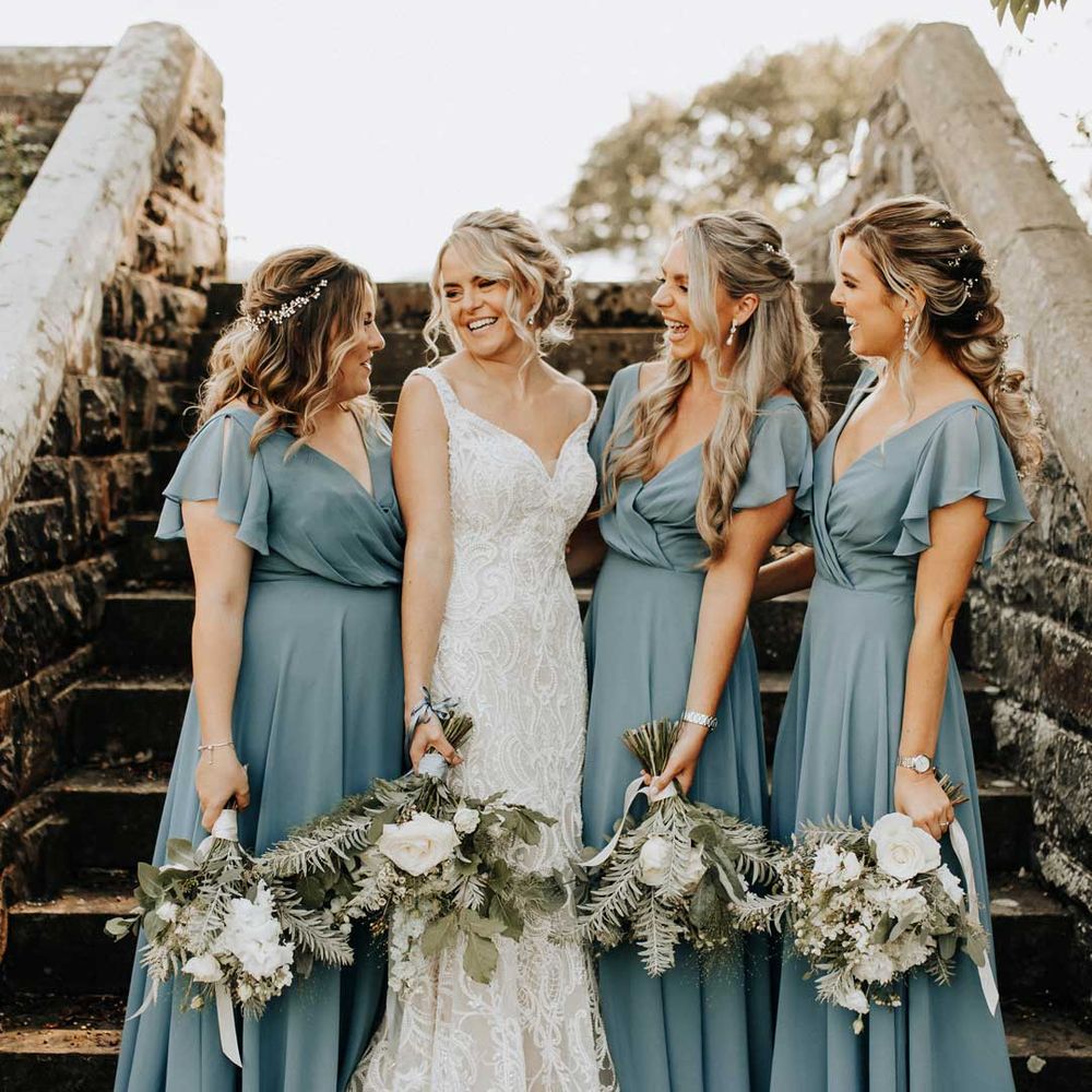 Bride in strappy lace wedding dress standing with bridal party wearing wrap front short sleeve blue bridesmaid dresses 