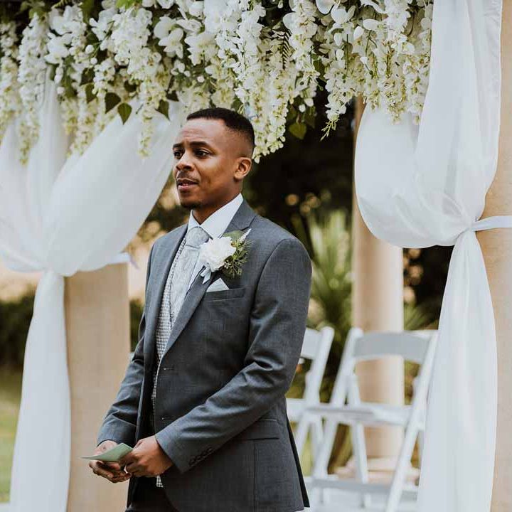 Groom in charcoal grey grooms suit, light grey patterned waistcoat, light grey patterned tie and large white rose and foliage boutonniere standing at the alter of Tredudwell Manor