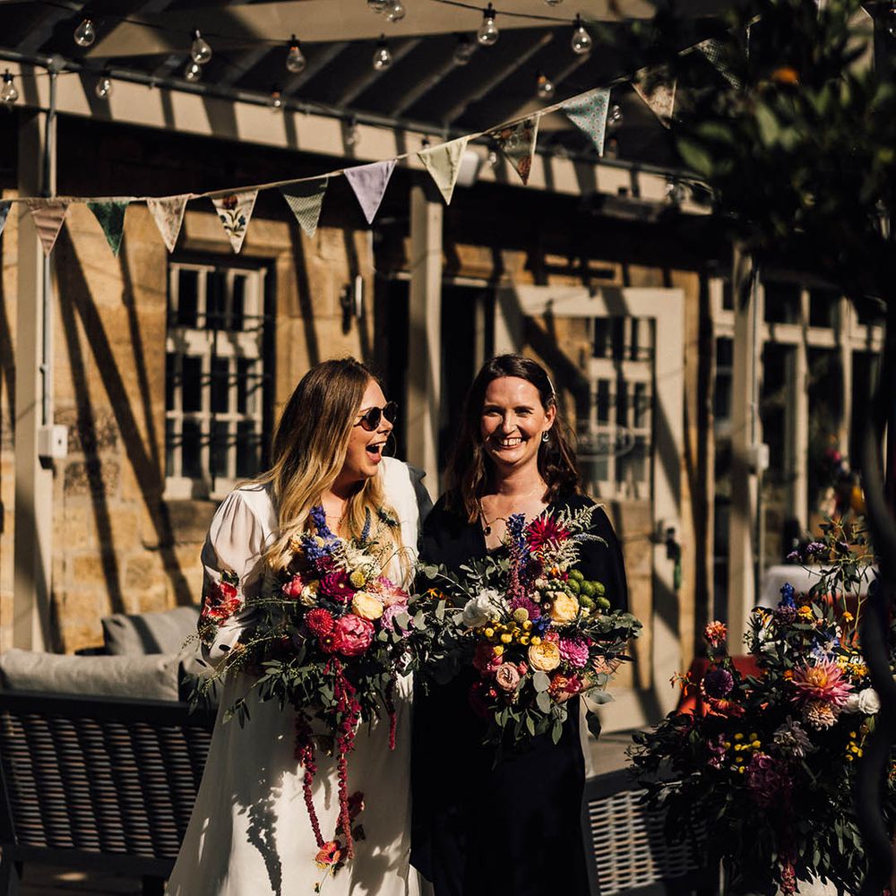 Bride in white wedding dress with colourful embroidered wedding flowers carrying a bouquet with the bride in a black wedding jumpsuit for lesbian wedding 