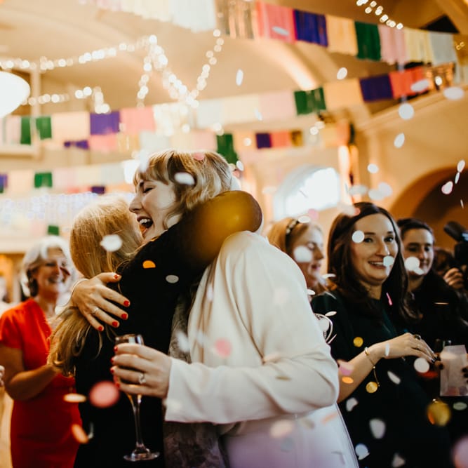 Bride in a long sleeve jumpsuit with red nail polish embracing a wedding guest during the confetti moment 