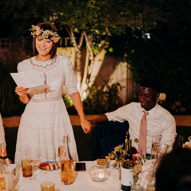 Bride in a white Broderie Anglaise dress with colourful embroidery ready her wedding speech at an outdoor reception 