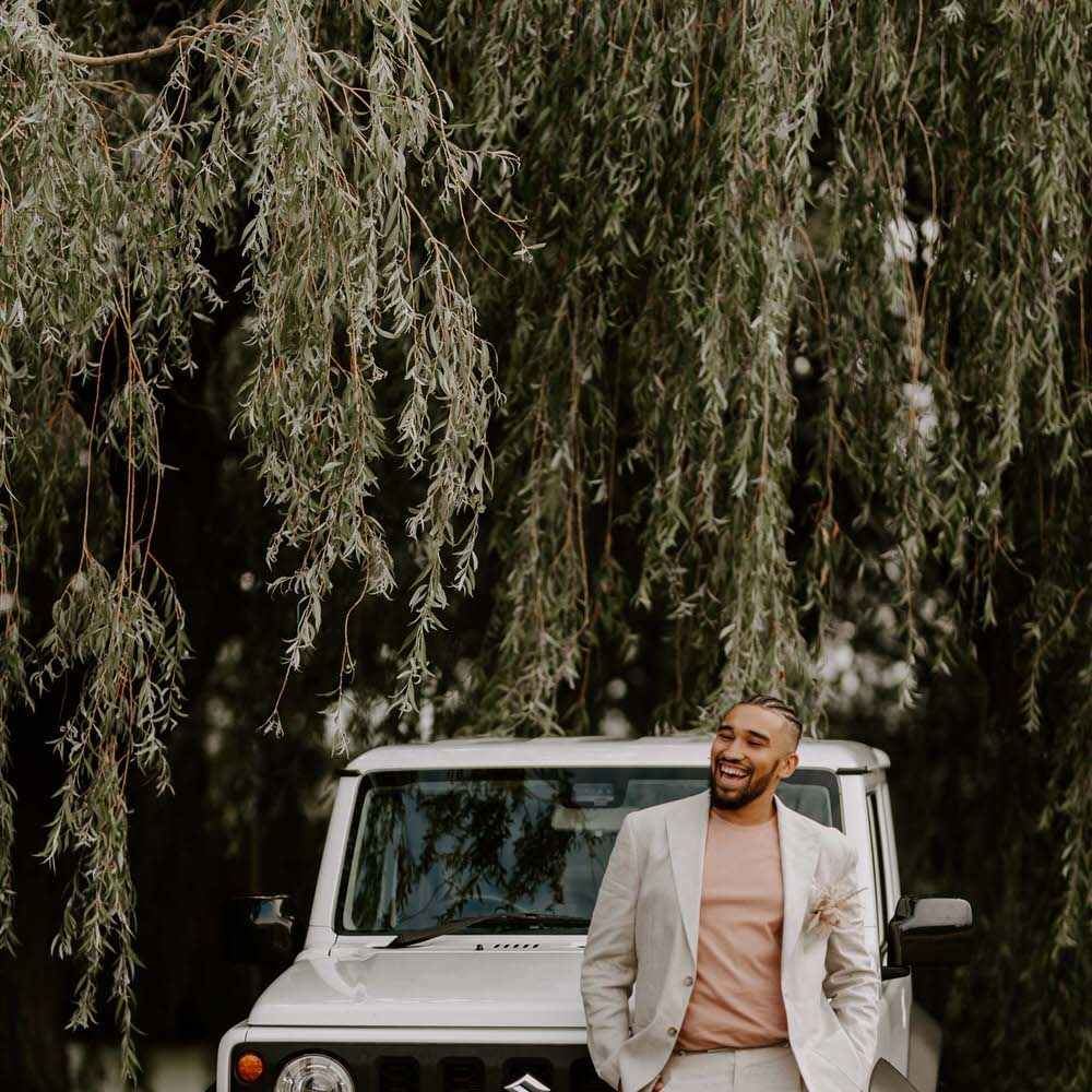 Groom in a cream suit leaning on a large white 4x4 Jeep