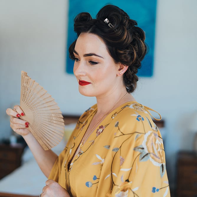 Bride has her hair pinned into curls on her wedding day as she fans her face and wears yellow dressing gown