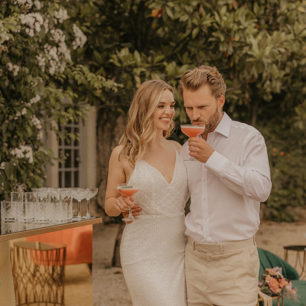 Groom in a white open collar shirt and beige trousers sipping cocktails with his bride in a sparkly Made With Love wedding dress
