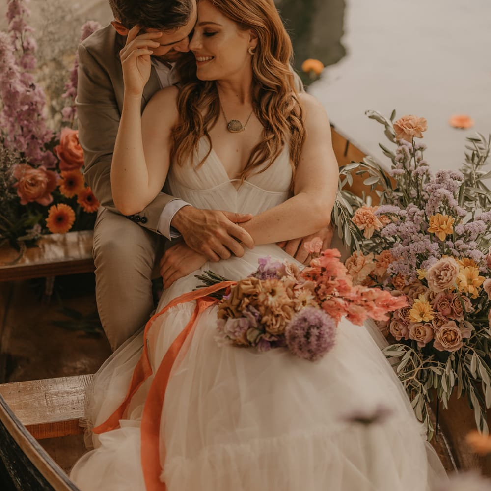 Groom in a beige suit embracing his bride in a Made With Love wedding dress as they sit on a boat at Euridge Manor surrounded by pastel wedding flowers 