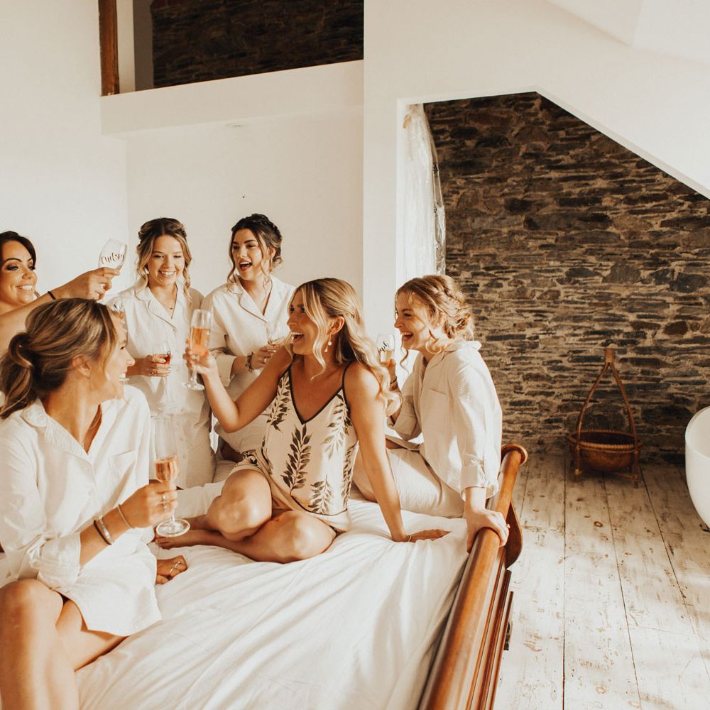 Bride in cami pyjama set and bridesmaids all in long white shirts raise flutes of sparkling wine as they sit on large bed before wedding at Anran Devon