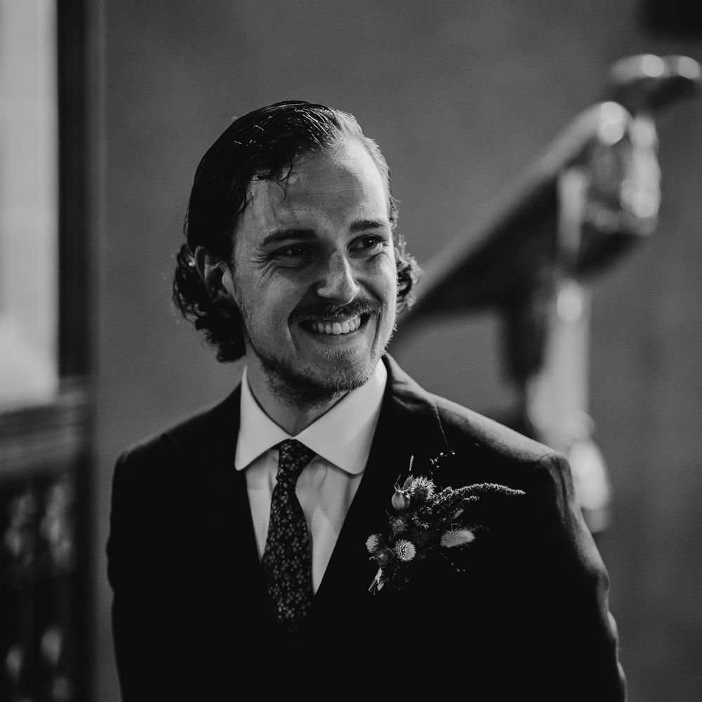 Groom in three piece suit, patterned tie and dried floral buttonhole smiles as he waits for brides during church wedding ceremony in Cornwall