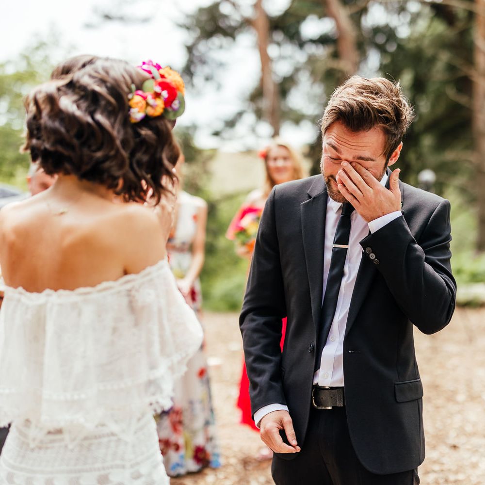 Groom in black suit wipes his eyes during outdoor wedding ceremony with bride in off the shoulder Grace Loves Lace wedding dress and colourful flower crown