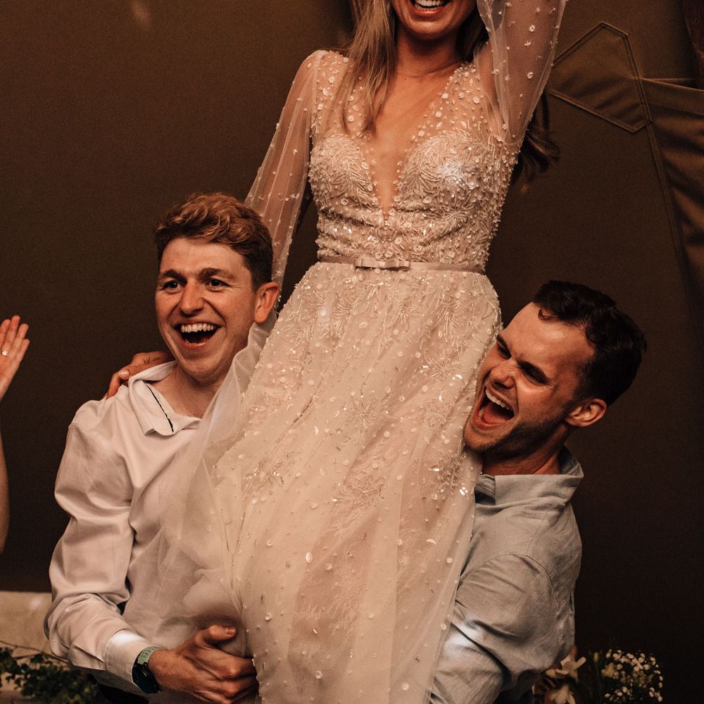 Bride in long mesh sleeve embroidered wedding dress and flower crown is held up on shoulders of two wedding guests at tipi wedding reception