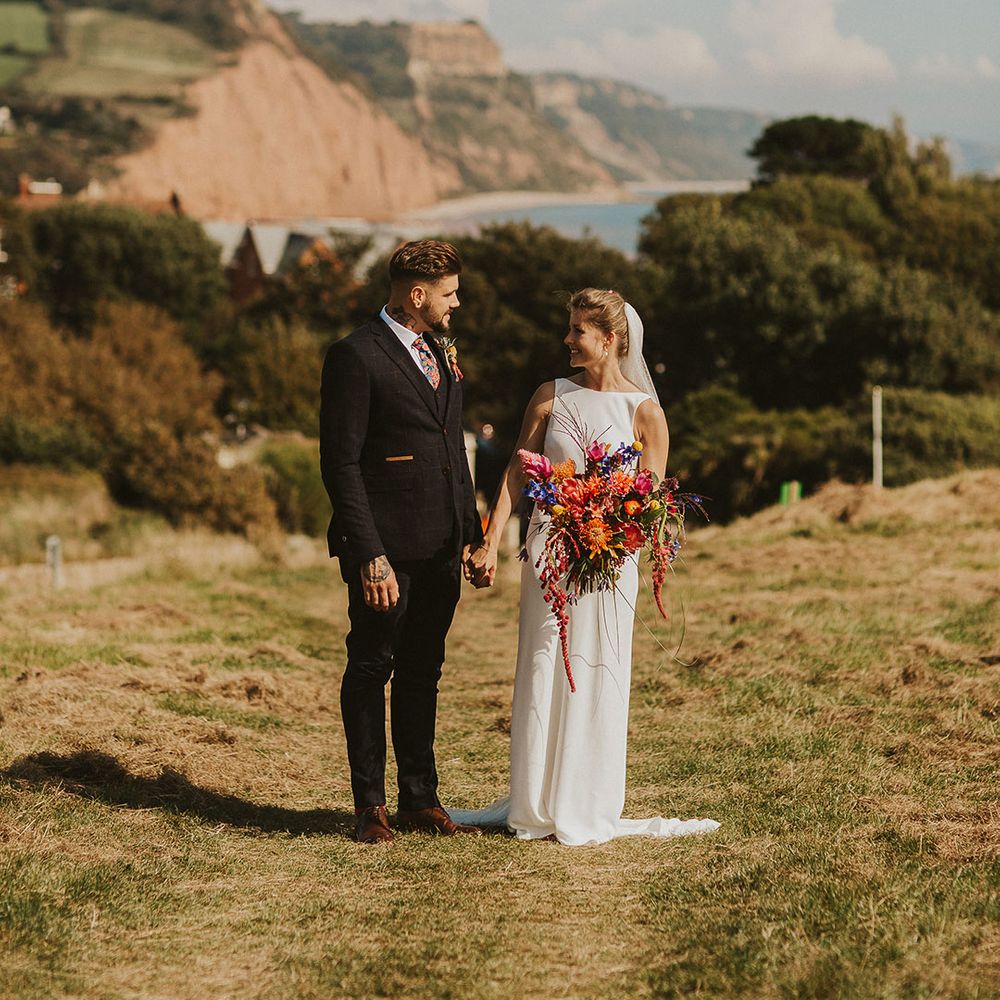 Coastal wedding with bride in Charlie Brear wedding dress and cathedral length veil holding a bright pink wedding bouquet and her grooms hand in a navy suit 