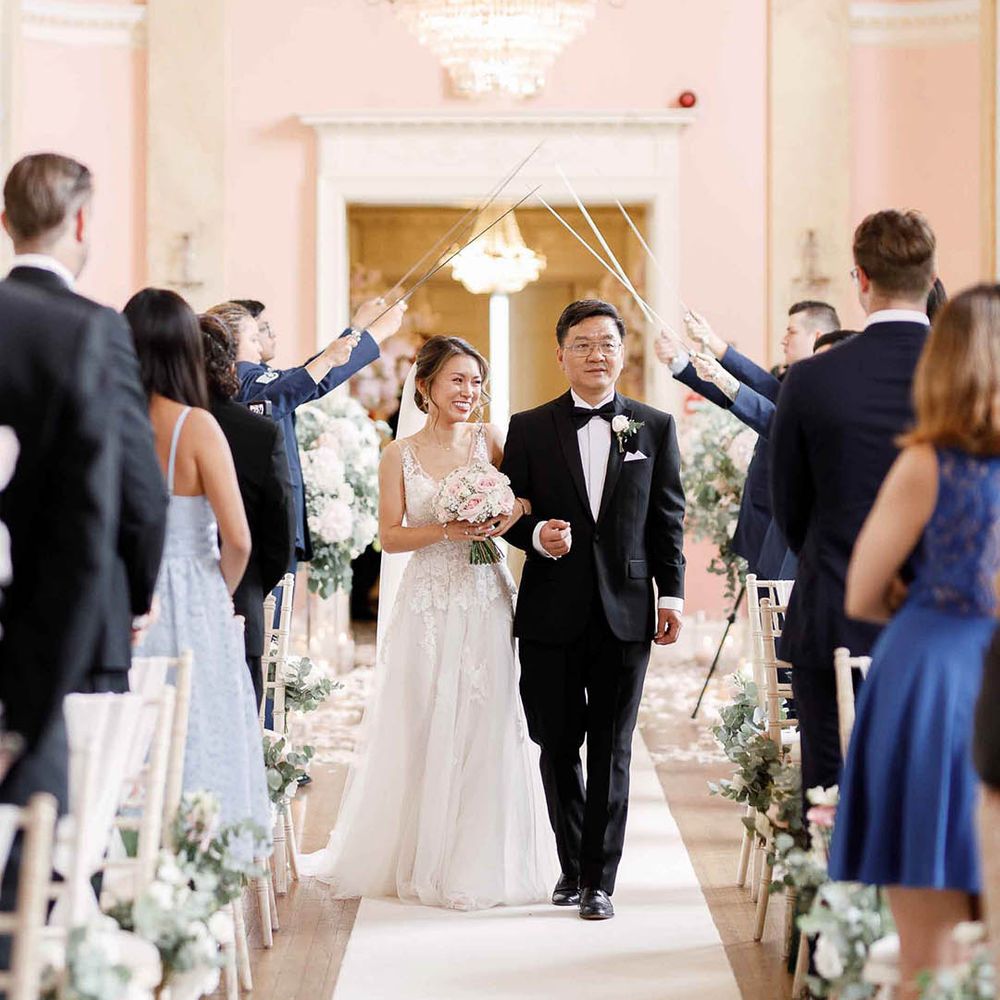 Bride walks down the aisle with her father who wears black tie as swords are held above them for Military ceremony