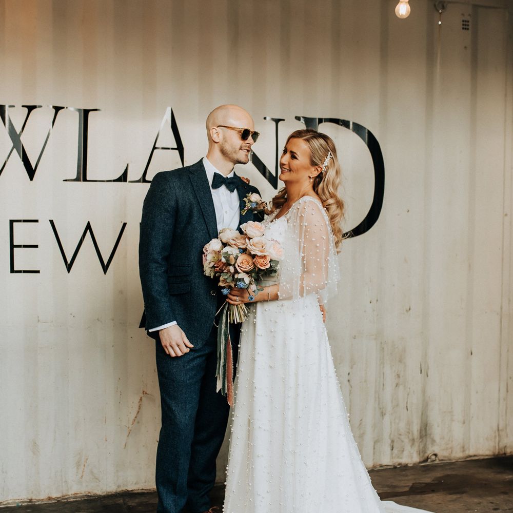 Bride in pearl wedding dress with sleeves and train holding pastel mixed rose wedding bouquet stands with groom in blue tweed suit with bow tie