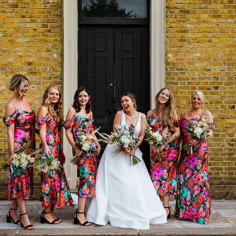 Bride stands with bridesmaids in bright floral gowns and black strappy heels in front of yellow brick wall in London