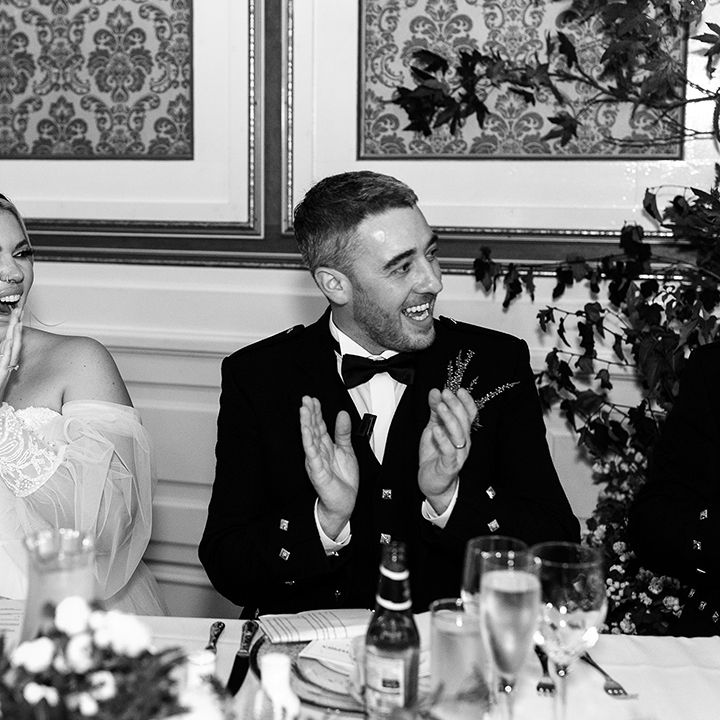 Bride in strapless wedding dress with sheer sleeves and lace cuffs and groom in black tie applaud for speeches
