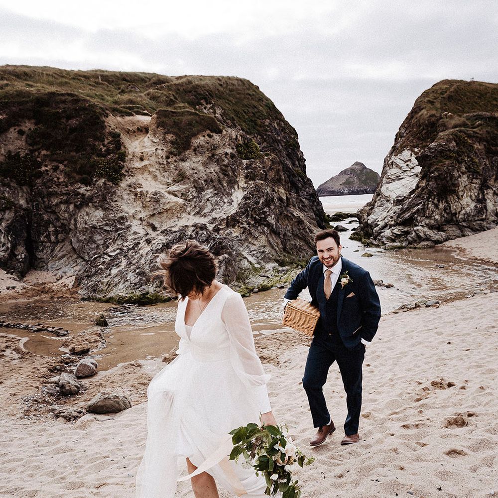 Bride walks barefoot in the sand in her Sassi Holford wedding dress as groom walks behind in blue suit carrying wicker picnic basket 