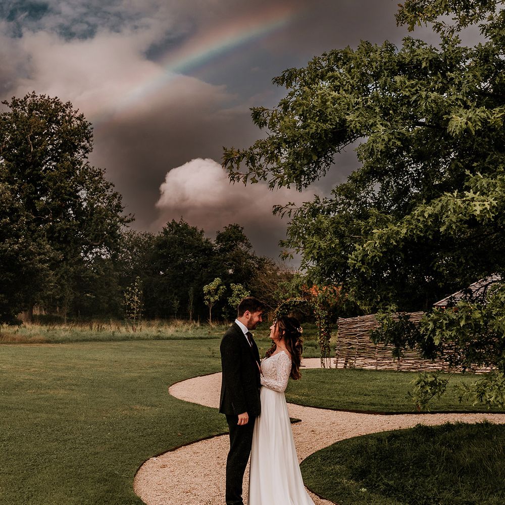 Bride places her hands on the groom's chest as they smile at each other underneath a cloudy sky and rainbow on their wedding day