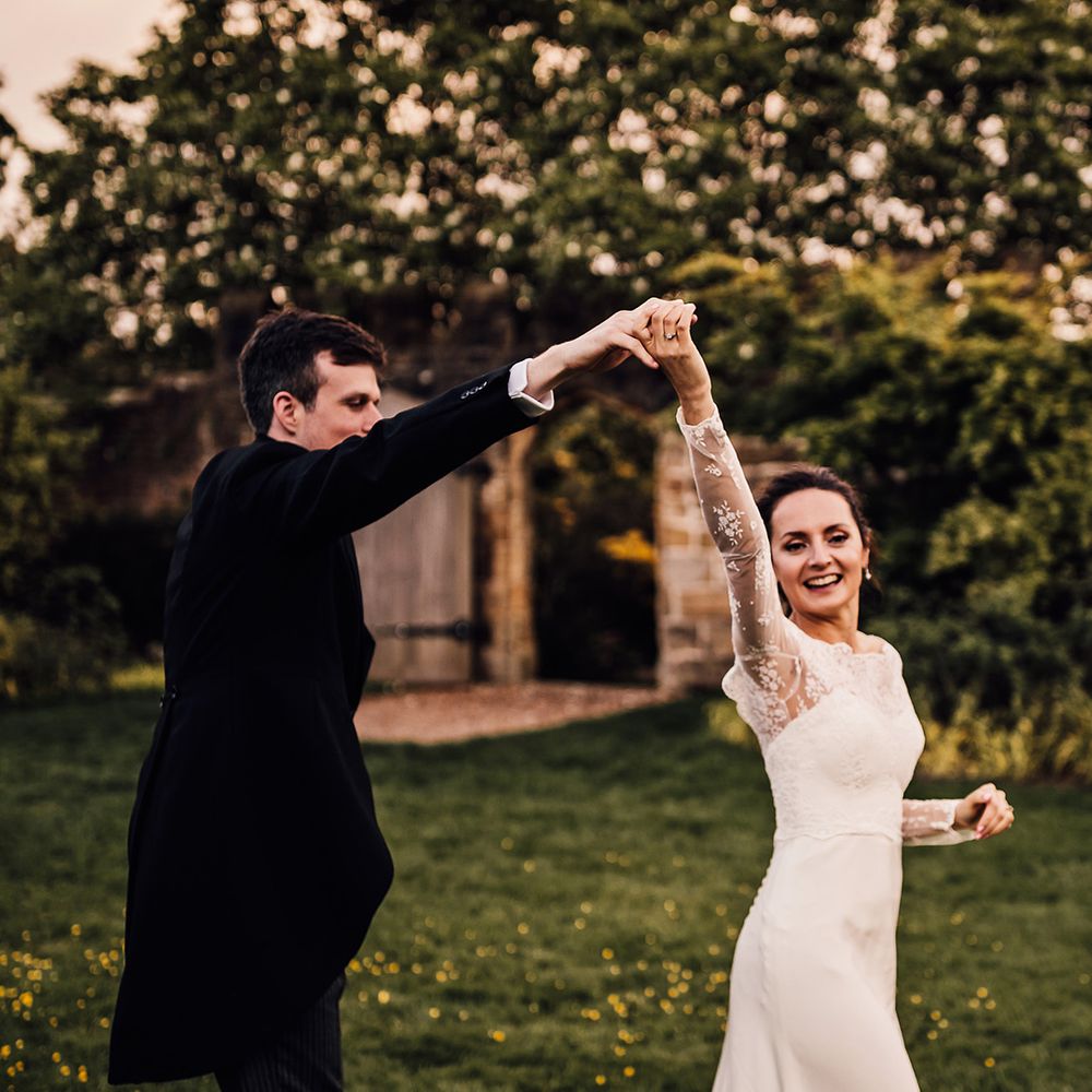Groom in black morning suit spins bride around in satin white wedding dress with sheer lace sleeves 