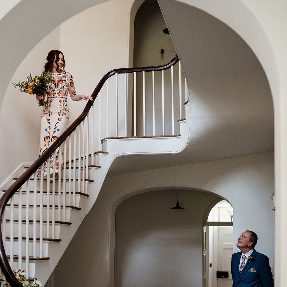 Father of the bride waits and looks up to get first look at the bride as she descends down the stairs in Temperley Bridal dress