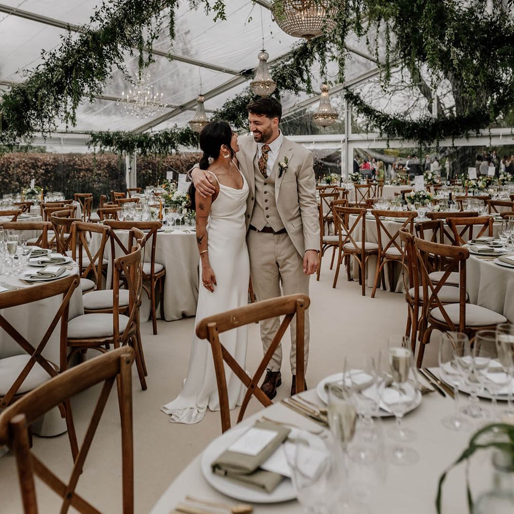 Bride in a satin wedding dress and groom in a beige suit standing in the middle of their clear glass marquee wedding reception with wooden furniture, greenery decor and handing chandelier 
