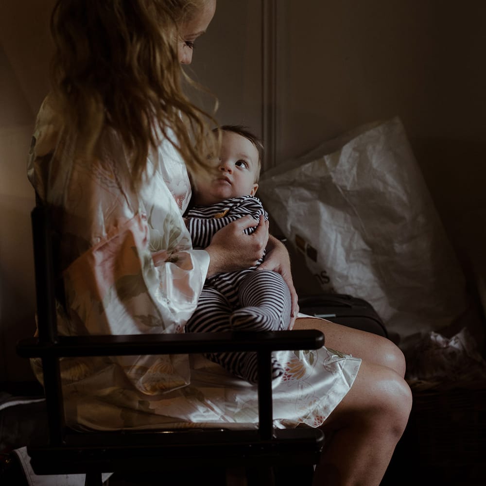 Bride in satin floral robe sits with baby in striped baby grow 