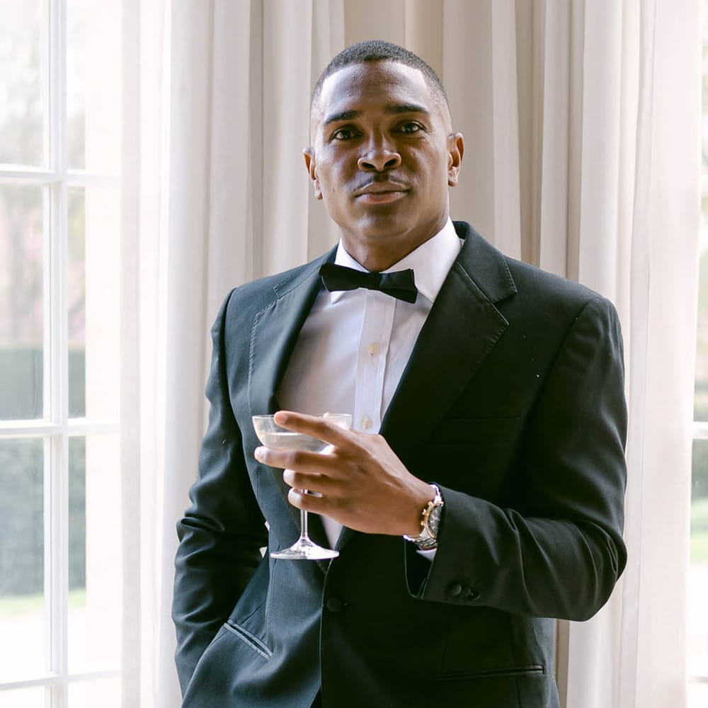 Groom stands looking at the camera in black tie with a cocktail glass 