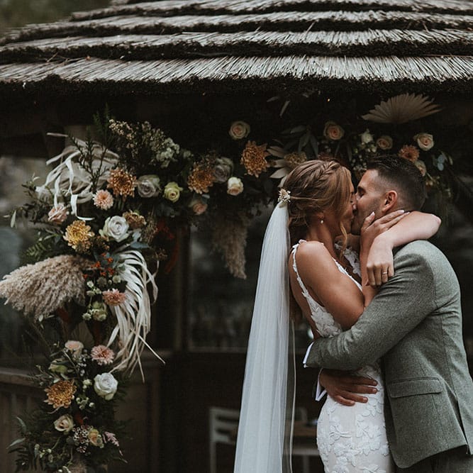 Bride & groom have their first kiss during wedding ceremony as champagne and rust florals line the front of the beach hut 