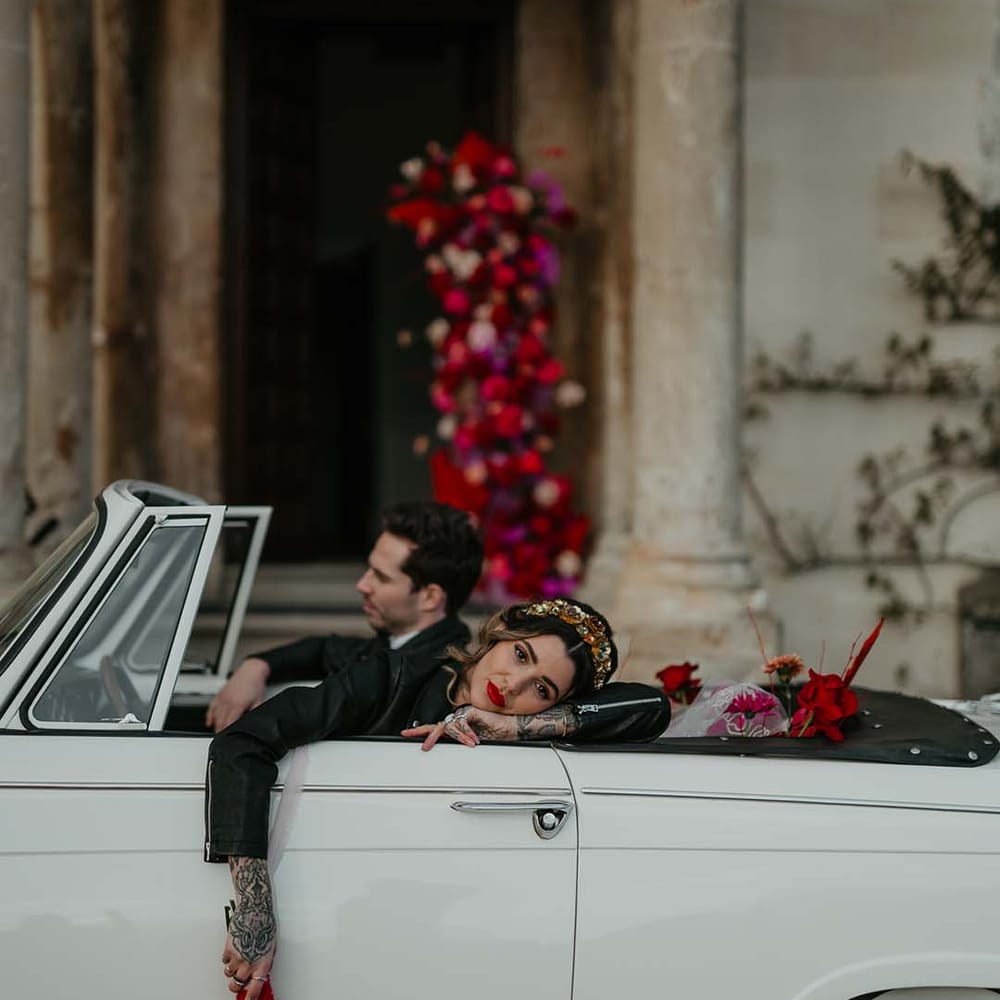 Bride leaning on off white classic convertible wedding car in leather jacket and large gold bridal headband holding hot pink, bright pink and magenta bridal bouquet with dahlias, orchids, roses, amaryllis, anthuriums and dried flowers 