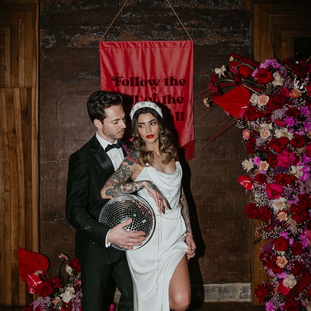 Bride in cowl neck sleeveless satin wedding dress with leg slit, gold glittery wedding headband, and soft grey fluffy feather open toe strappy high heels and groom in black tuxedo with magenta amaryllis and orchid boutonniere posing with disco ball wedding decor