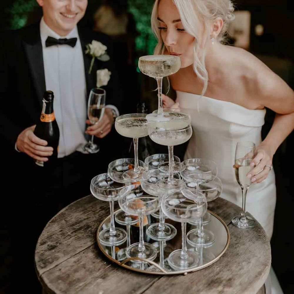 Bride in strapless satin wedding dress sipping from wedding champagne tower with groom in classic black tuxedo 