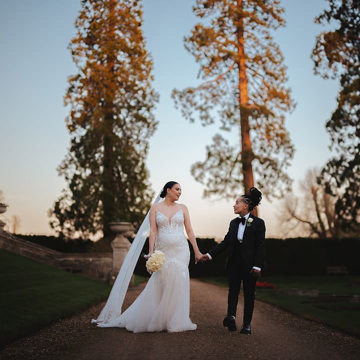 Bride in lace sleeveless v neck Enzoani wedding dress with tulle finish and church length veil walking hand in hand with bride in black snakeskin print tuxedo with black bowtie and white rose boutonniere walking through the grounds of Grittleton House wedding venue 