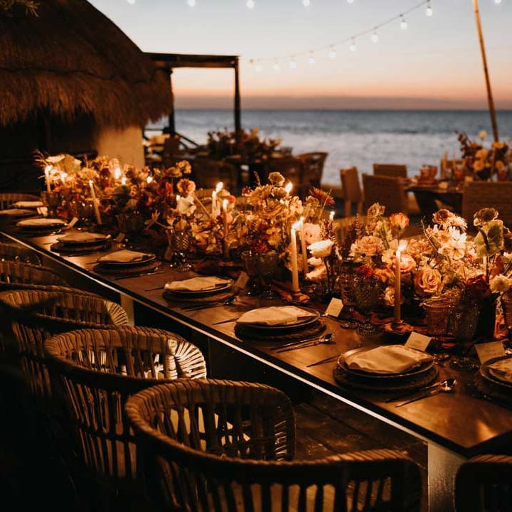 Rustic wedding tablescape by the Mexican sea with wicker chairs, burnt orange dried roses, dried flower and foliage floral centrepieces, wooden slate wedding table numbers, light orange tapered candles and burnt orange table runners under suspended fairy light wedding lighting 