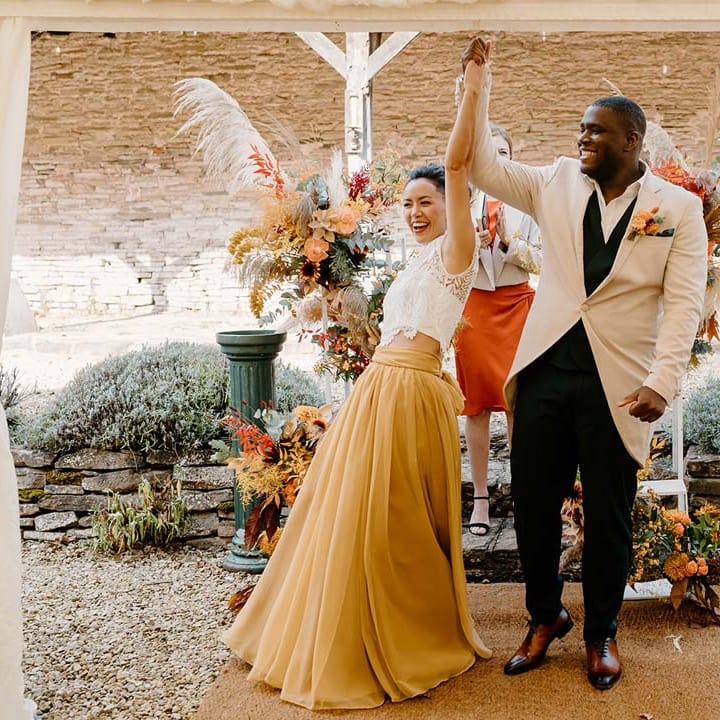 Bride in white mesh lace bridal crop top and yellow wedding skirt holding hands and cheering with groom in cream morning suit with forest green grooms waistcoat and trousers, forest green pocket square and orange boutonniere 