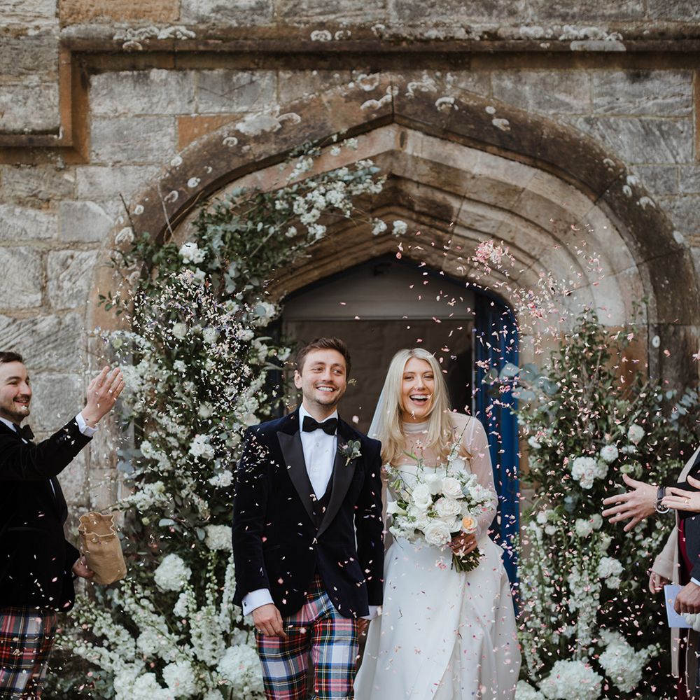 White wedding flower columns decorating the church entrance with the groom in tartan trousers and bride in mesh long sleeve wedding dress as guests throw confetti