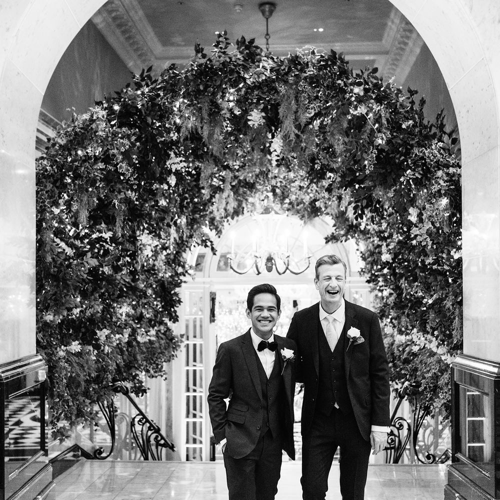 Black & white image of grooms walking through floral archway 