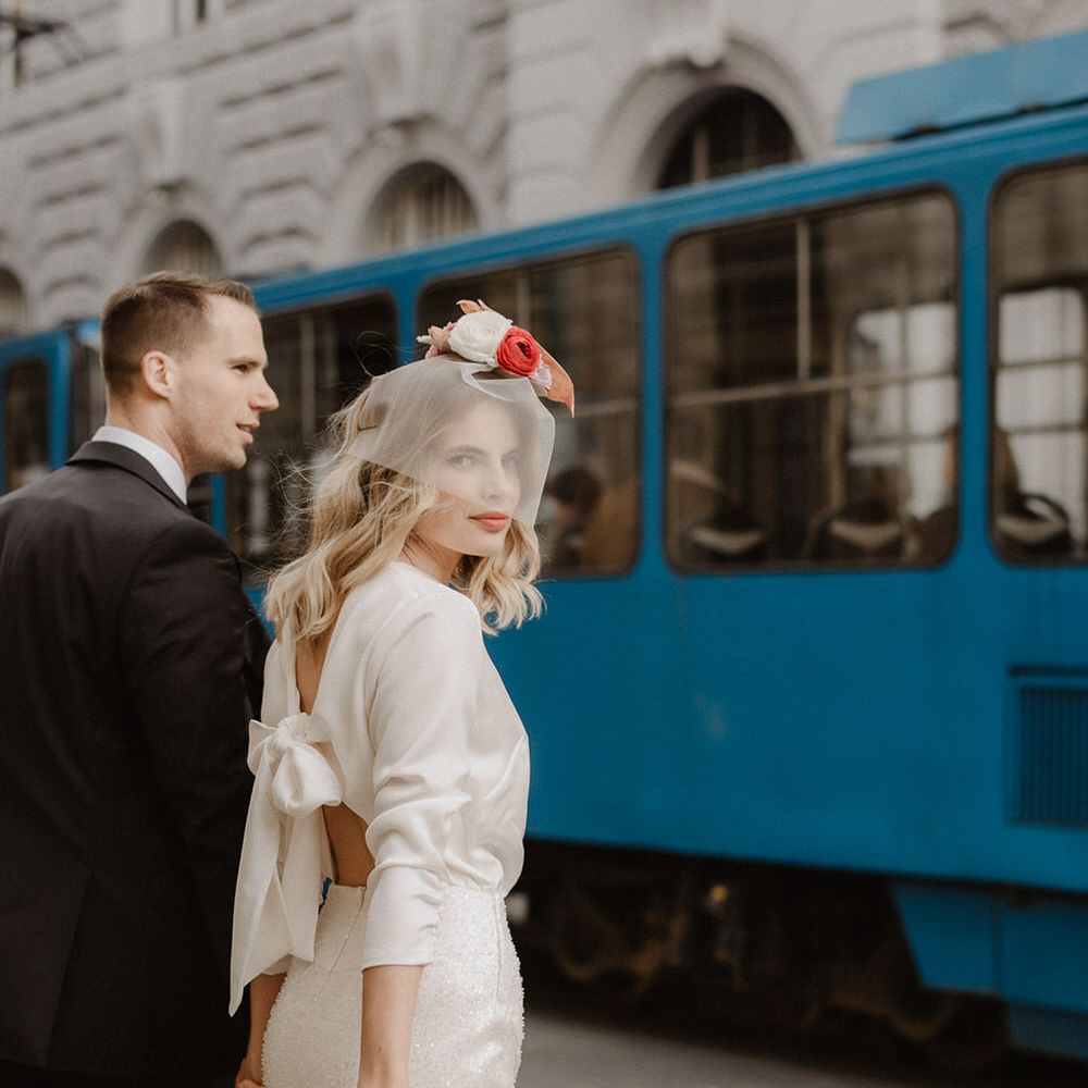 Bride and groom in front of a tram in the city, the bride wearing an open back wedding dress by Gray Bridal with bow detail. Holding a bouquet of white and pink flowers